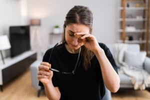 Picture of a woman taking her glasses off and touching her eye in pain because she needs pink eye treatment.